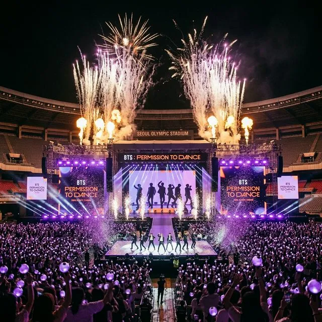 Palco do concerto de comeback do BTS no Estádio Olímpico de Seul, com fogos de artifício e luzes púrpura iluminando 100 mil fãs